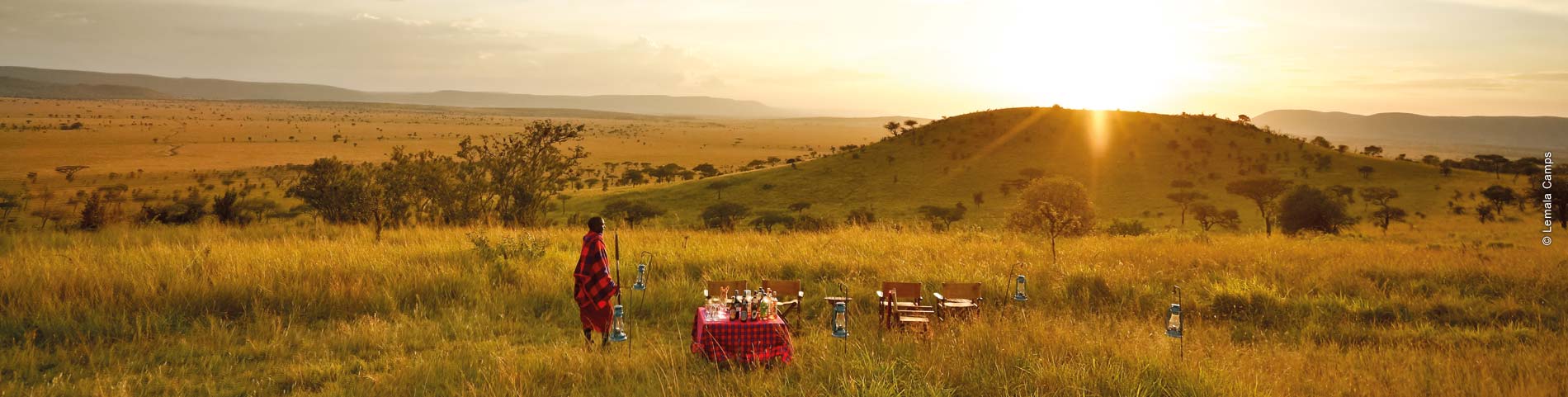 Person steht in einem weitläufigen mit Gras bewachsenen Gebiet mit kleinem Hügel. Daneben steht ein bedeckter Tisch mit Sitzmöglichkeiten.