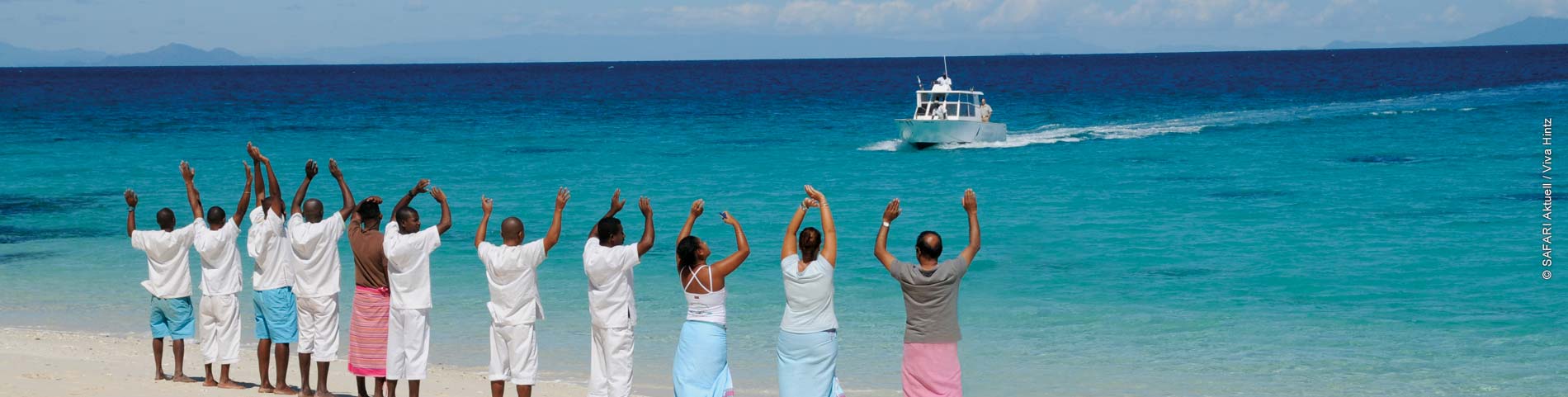 Gruppe Menschen jubelt auf einem Strand und im Hintergrund fährt ein Boot im blauen Meer