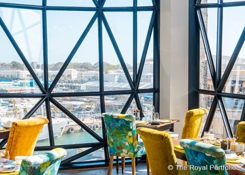 Restaurant mit bunten Stühlen vor großen, gitterförmigen Panoramafenstern und Blick auf den Hafen.