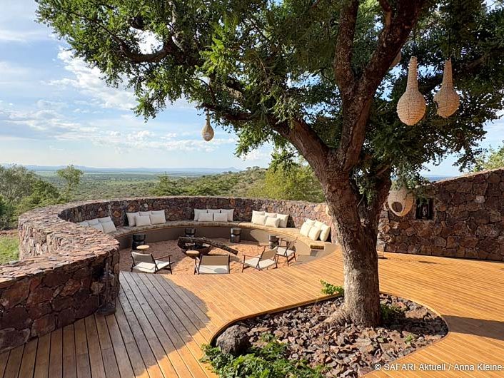 Terrasse mit Sitzgelegenheiten und einer sonnigen Aussicht auf die afrikanische Landschaft. Im Vordergrund ein großer Baum mit kleinen Vogelkörben.