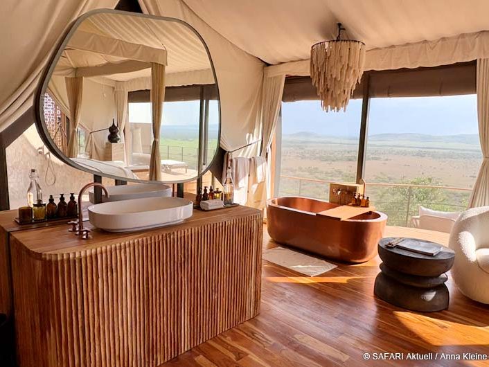 Badezimmer mit Waschbecken und großem Spiegel auf einer Holzkommode. Im Hintergrund Badewanne und große Fenster mit weitem Ausblick.