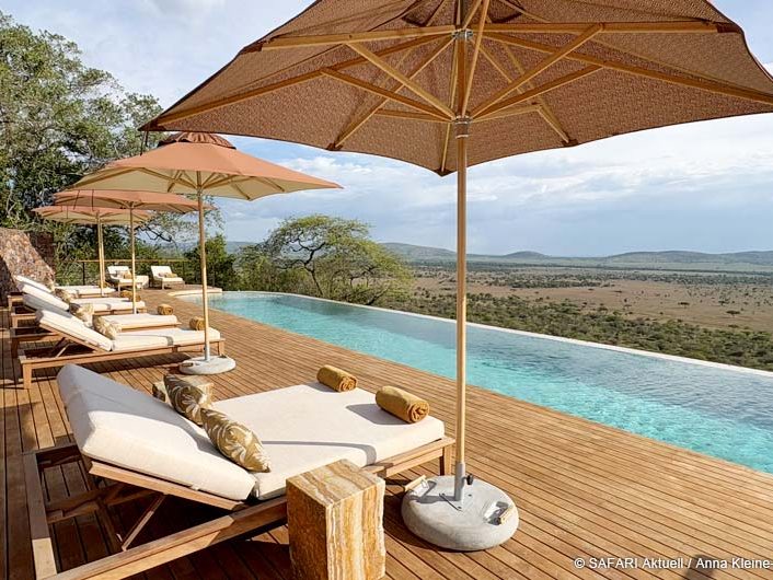 Im Vordergrund mehrere Liegestühle und Sonnenschirme. Dahinter ein langgezogener Pool mit Ausblick auf die afrikanische Landschaft.